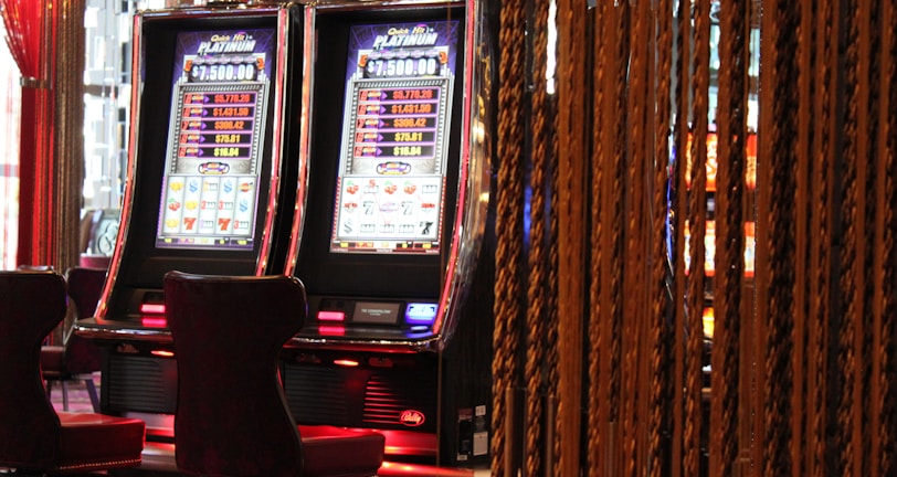 A pair of casino slot machines, each with detailed screens displaying various game outcomes and jackpot amounts. The chairs in front of the slot machines are dark and cushioned for comfort. The background features a hanging curtain made of golden ropes and other casino decorations are visible.