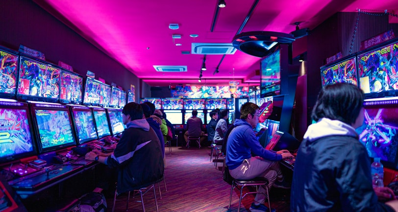 A vibrant arcade with rows of gaming machines adorned with colorful screens. Several people are immersed in playing games, seated on stools in a dimly lit room accented by an intense pink ceiling light. The atmosphere is lively and engaging.