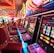A brightly lit casino interior featuring a row of colorful slot machines. The neon lights create a vibrant atmosphere, with patterned carpeting adding to the decor. Stools are aligned neatly in front of each machine.
