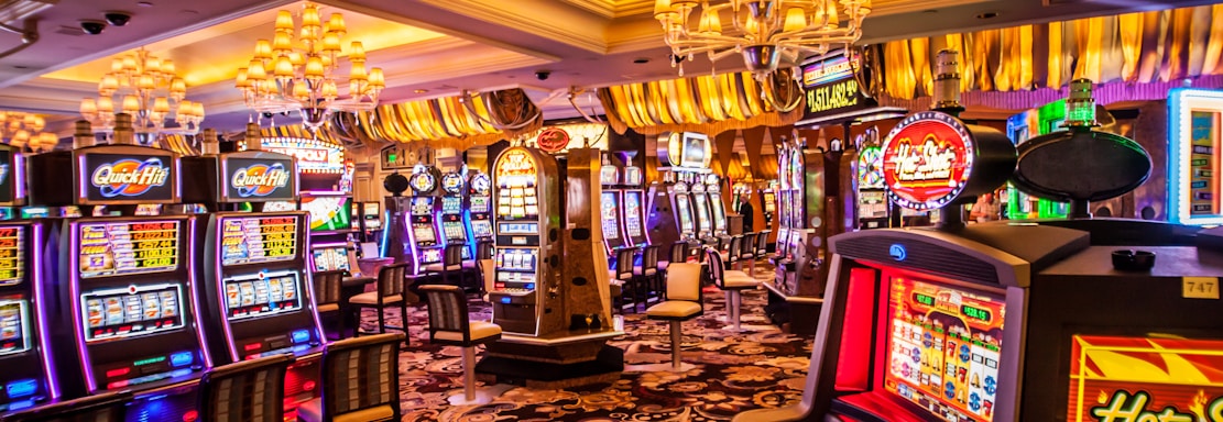 A vibrant casino interior filled with rows of colorful slot machines, each illuminated with bright lights and displays. The ceiling is adorned with elegant chandeliers and golden drapes, creating a luxurious atmosphere. The patterned carpet adds to the elaborate design of the space.