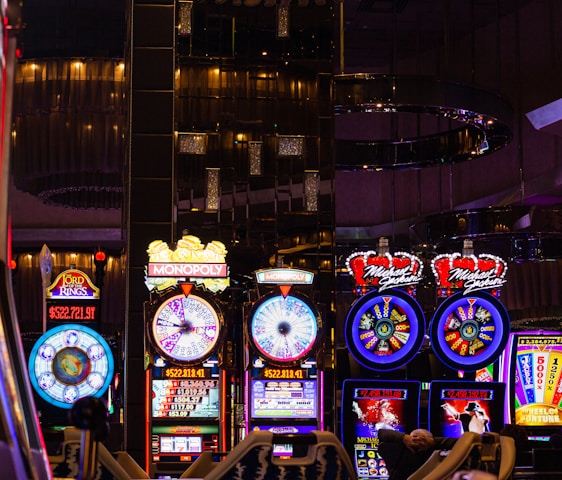 A lively casino scene featuring a row of bright, colorful slot machines with themes like Monopoly, Michael Jackson, and Lord of the Rings. The gaming machines display a variety of flashing lights and digital screens, creating an energetic and exciting atmosphere. Reflections of the lights are visible in glossy surfaces above the machines.