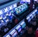 A bustling arcade filled with rows of gaming machines, each with vibrant displays and controls. Several people are seated at these machines, deeply engaged in playing games. Bright fluorescent lights cast a blue and neon glow across the room, enhancing the lively and energetic atmosphere.